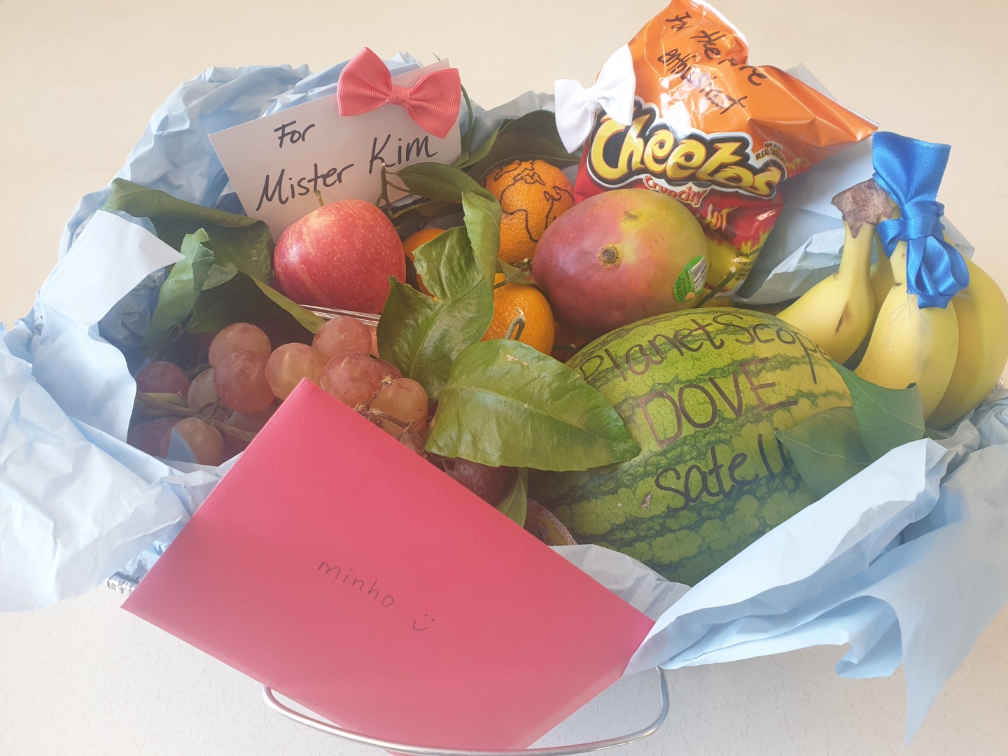 A fruit basket gifted by students, labeled with remote-sensing in-jokes: a watermelon marked 'PlanetScope DOVE Satellite' and a card reading 'For Mister Kim'
